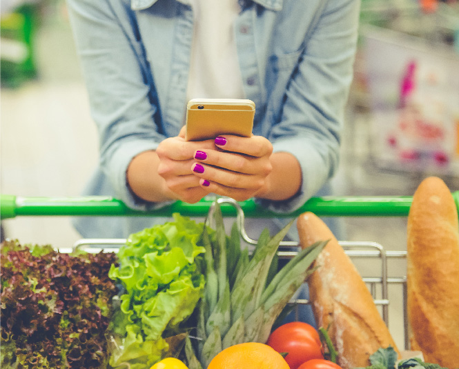 Young woman with a grocery cart full of produce holding her phone.
