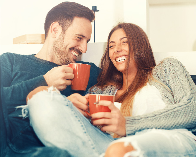 Young couple on a couch smiling and holding coffee mugs.